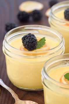 four small jars filled with pudding and topped with blackberries on a wooden table next to two spoons