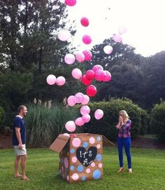 a man and woman standing in the grass near a cardboard box with balloons floating from it