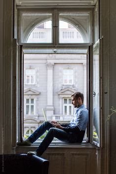 a man sitting on a window sill using a laptop computer by an open window