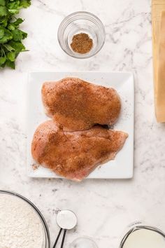 two pieces of chicken on a white plate next to some ingredients and utensils