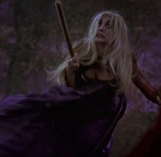 a woman with long blonde hair holding a wooden stick in her hand and wearing a purple dress