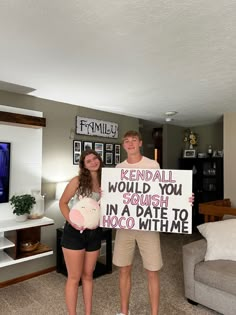 a man and woman standing in a living room holding a sign that says, rendal would you crush in a date to hoo witme?