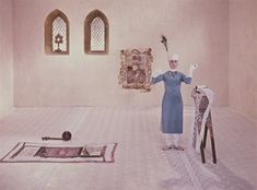 a woman standing in a room holding two brooms and wearing a white hat with an ornate wall hanging behind her