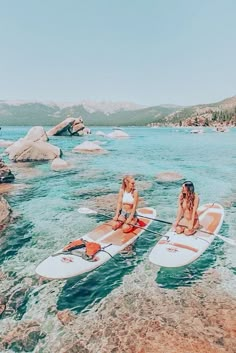 two women are sitting on surfboards in the water near rocks and boulders, while another woman is standing behind them