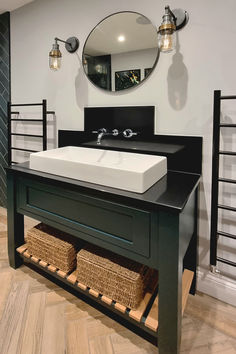 A bathroom with industrial wall lights, a round mirror, a black vanity with a white rectangular sink, and wicker storage baskets beneath. Warm Wood Flooring, Sophisticated Bathroom, Wicker Storage, Bespoke Kitchen, Black Vanity, Wicker Baskets Storage, Industrial Wall, Bespoke Kitchens, Filament Bulb