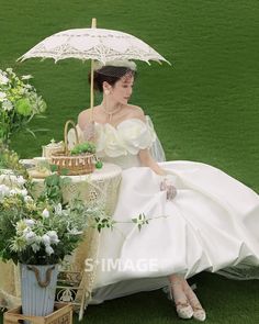 a woman in a wedding dress sitting under an umbrella on the grass next to flowers