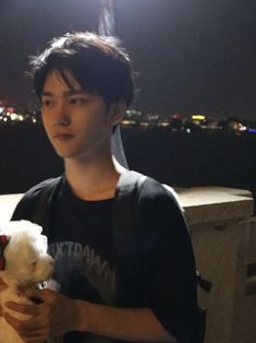 a young man holding a teddy bear in his hands at night time with the city lights behind him