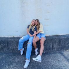 two young women sitting on the side of a building together, posing for a photo