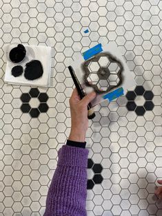 a person is drawing on the floor with black and white tiles in the background, while another hand holds a pencil