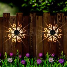 two lights that are on the side of a wooden fence in front of some flowers