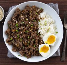 a white plate topped with meat and rice next to an egg on top of a wooden table