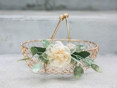 a white flower in a basket with green leaves on the side and a gold handle