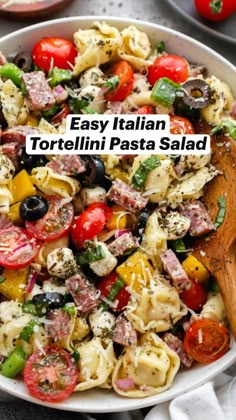 a pasta salad with tomatoes, olives and other ingredients in a white bowl next to a wooden spoon