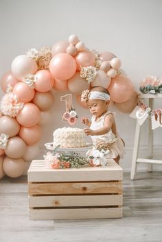 a baby girl standing in front of a cake with balloons on the wall behind her