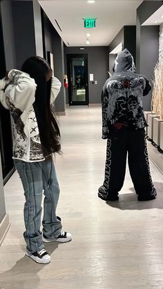 two people are standing in the hallway looking at their cell phones and one person is wearing a hoodie