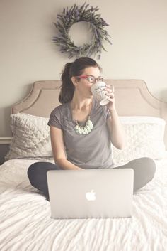 a woman is sitting on her bed drinking coffee and working on her laptop