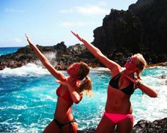 two women in bikinis jumping into the air on rocks by the ocean with blue water