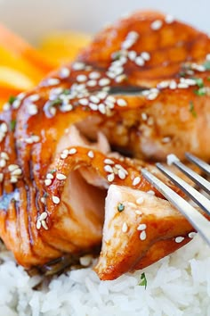 two pieces of salmon on top of white rice with sesame seeds and carrots in the background