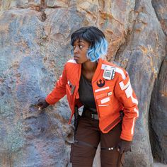 a woman with blue hair is standing in front of a rock wall wearing an orange jacket and brown pants