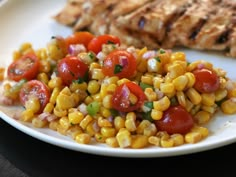 a white plate topped with corn and tomatoes next to grilled meat on a table