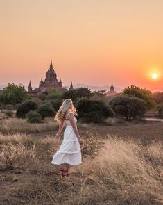 a woman in a white dress is walking through the grass at sunset with an old building in the background