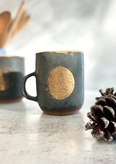 two coffee mugs sitting on top of a counter next to a pinecone