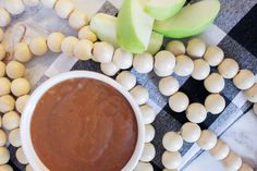 a bowl of peanut butter next to some peanuts and an apple on a checkered tablecloth