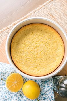 a lemon cake in a white bowl on a blue and white cloth next to two lemons