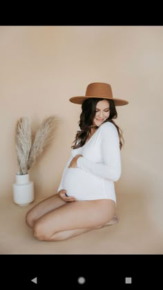 a pregnant woman sitting on the ground wearing a brown hat and white bodysuit with her legs crossed