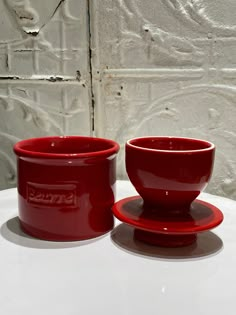 two red cups and saucers sitting on top of a white table next to each other