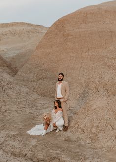 a man and woman standing next to each other in front of some hills with sand