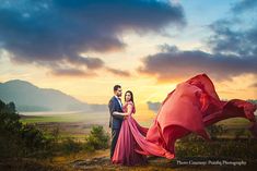 a man and woman standing on top of a hill next to each other in front of a red dress