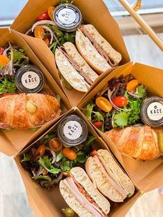 four boxes filled with sandwiches and salads on top of a wooden table in front of a window