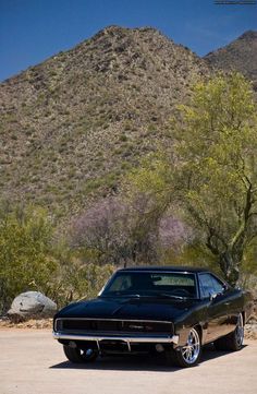 a black muscle car parked in front of a mountain with trees and bushes behind it