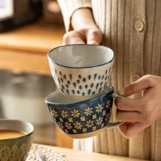 a woman is holding three cups in her hands while sitting at a table with two coffee mugs