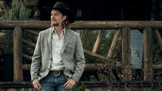 a man wearing a cowboy hat standing in front of a log cabin with his hands on his hips