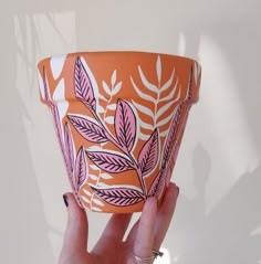 a hand holding an orange and pink pot with leaves painted on the outside, in front of a white wall