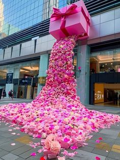 a large pink gift box is surrounded by flowers and petals on the ground in front of a building