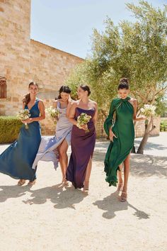 four bridesmaids walking in the sand with their dresses draped around them and holding bouquets