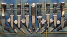 seven knives are lined up in a row on a plaid cloth, one is silver and the other is black