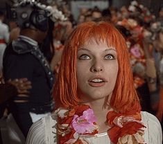 a woman with red hair and flowers around her neck standing in front of a crowd