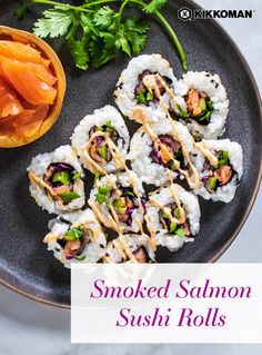 Image shows a dark gray plate, filling almost the entire frame, loaded with 10 sushi bites filled with smoked salmon, cucumber, purple cabbage, and cilantro. The rolls are drizzled with sriracha mayo. Also on the plate is a little wooden bowl with more smoked salmon, and the plate is garnished with a sprig of cilantro. The Kikkoman logo is at the top right of the image and the text overlay reads "Smoked Salmon Sushi Rolls" in a purple font on a transparent white background. Salmon Cream Cheese Sushi Roll, Sushi With Smoked Salmon, Smoked Salmon Sushi Recipes, Cooked Salmon Sushi Rolls, At Home Sushi Recipes, Smoked Salmon Sushi Roll, Smoked Salmon Rolls, Salmon Sushi Recipes, Easy Sushi Rolls