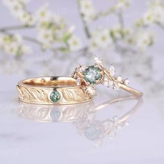 two gold wedding rings with green and white stones on the side, one is in front of flowers