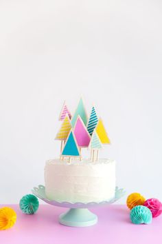 a white cake topped with colorful paper trees on top of a pink table next to cupcakes