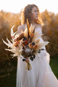 a woman in a white dress holding a bouquet with dried flowers and feathers on it