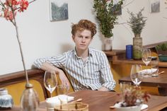 a young man sitting at a table with wine glasses and flowers in vases behind him