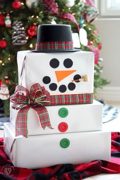 a stack of wrapped presents with a snowman on top and a christmas tree in the background