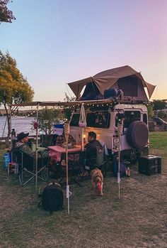 a camper van parked next to a lake with people sitting in the shade under it