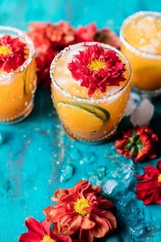 three glasses filled with drinks sitting on top of a blue table next to flowers and petals