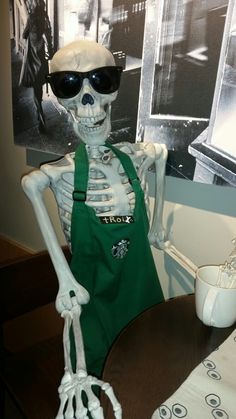 a skeleton in an apron and sunglasses sitting at a table with a cup on it